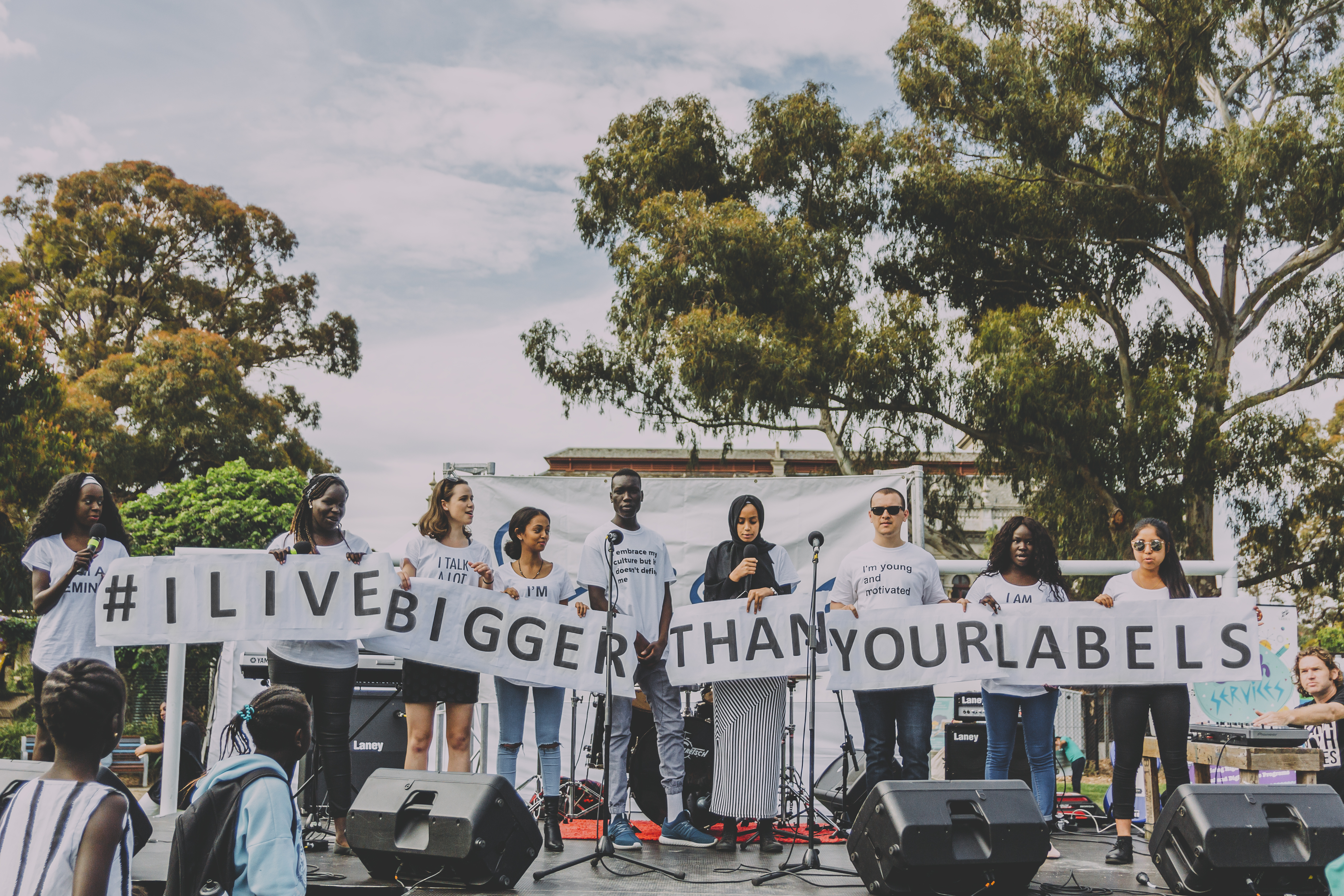 A group of young people on an outdoor stage holding signs that read I live bigger than your labels
