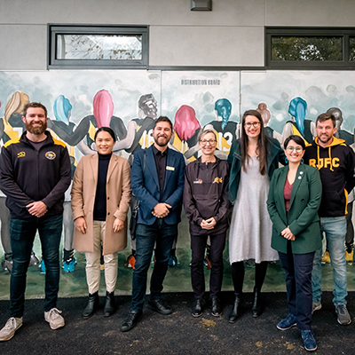 A group of men and women representing Richmond sporting clubs and Yarra City Council stand in front of a mural that pictures a women's football team standing arm in arm with their backs turned.