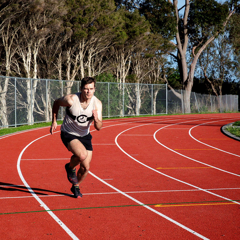 A man running on an athletics track with a fence and trees in the background