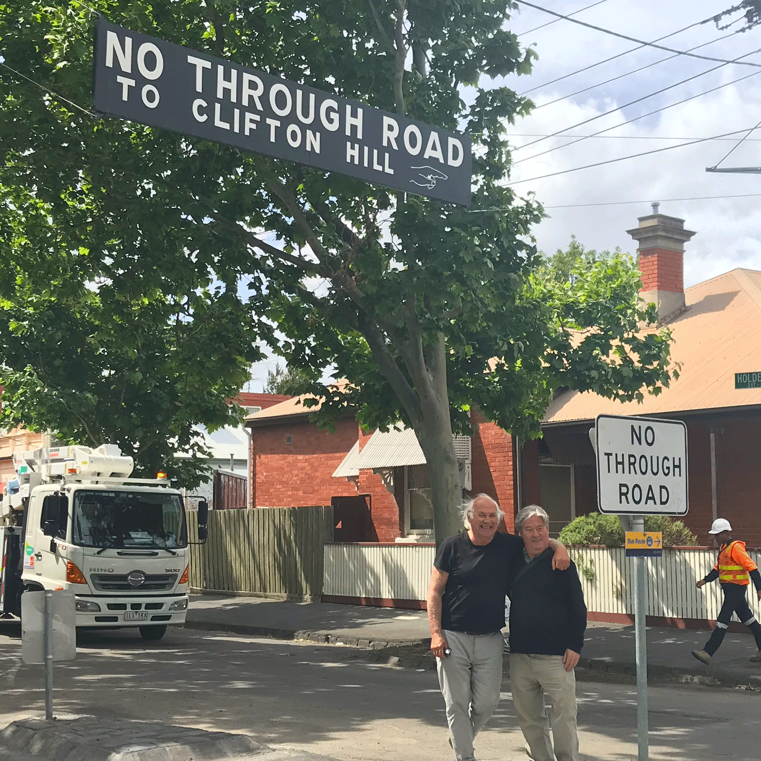 Two men standing arm in arm smiling below a No through road sign