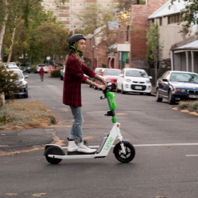 White and green electric scooter, driving down residential road by person in jeans, white boots and a plaid jacket. Cars parked in background.