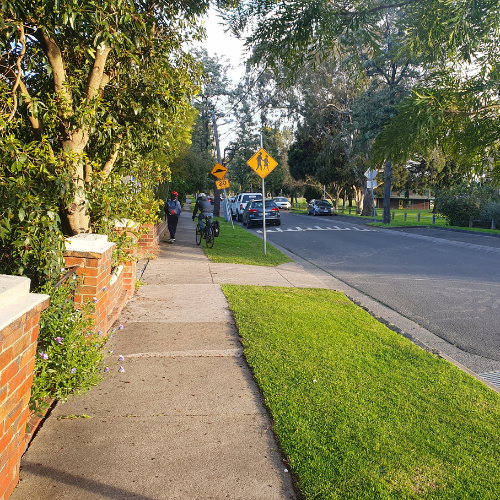 Photo of street in Alphington, showing a footpath with pedestrians, grass verge and road, with cars.