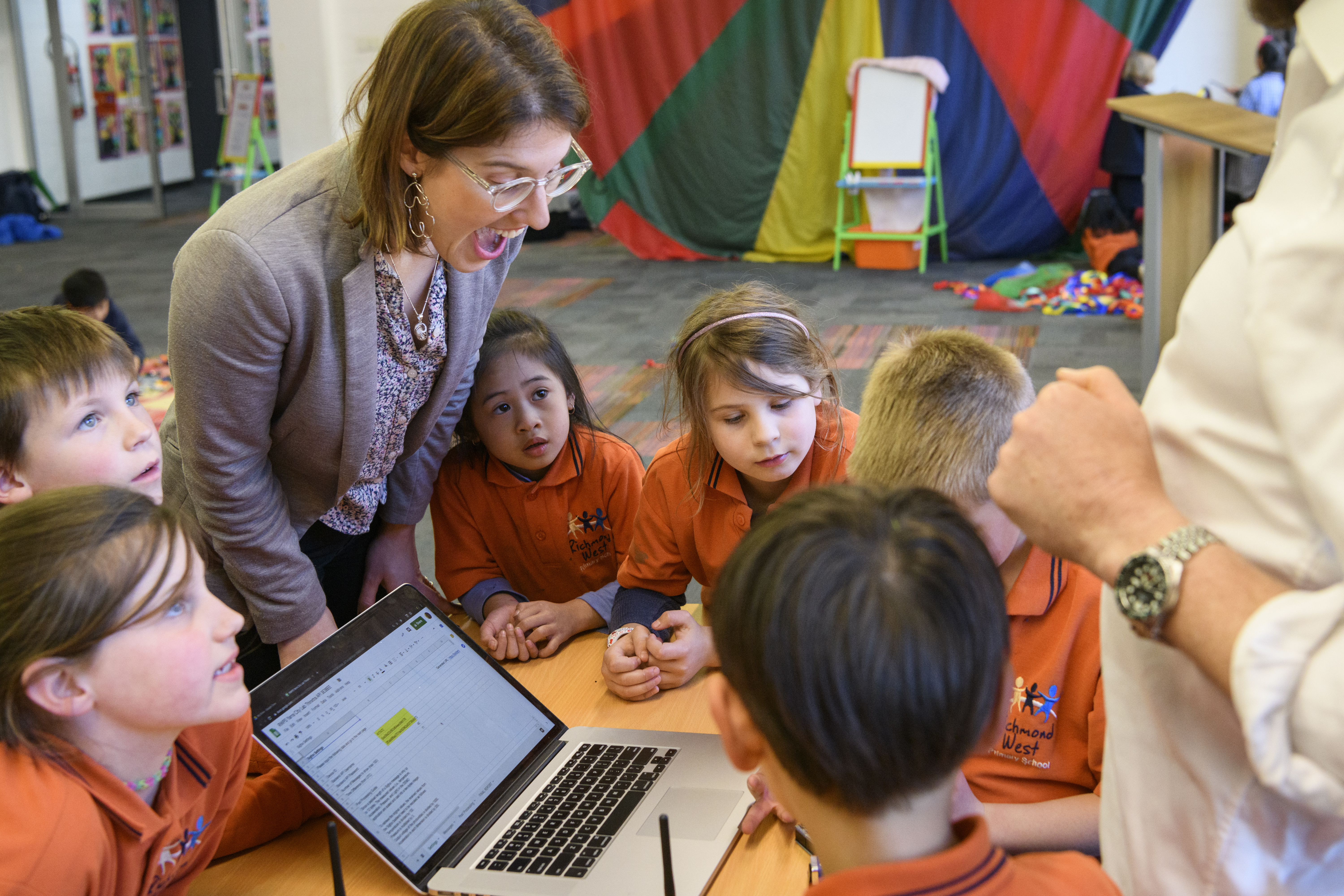 Some children in a school uniform and a young woman gather around a laptop.