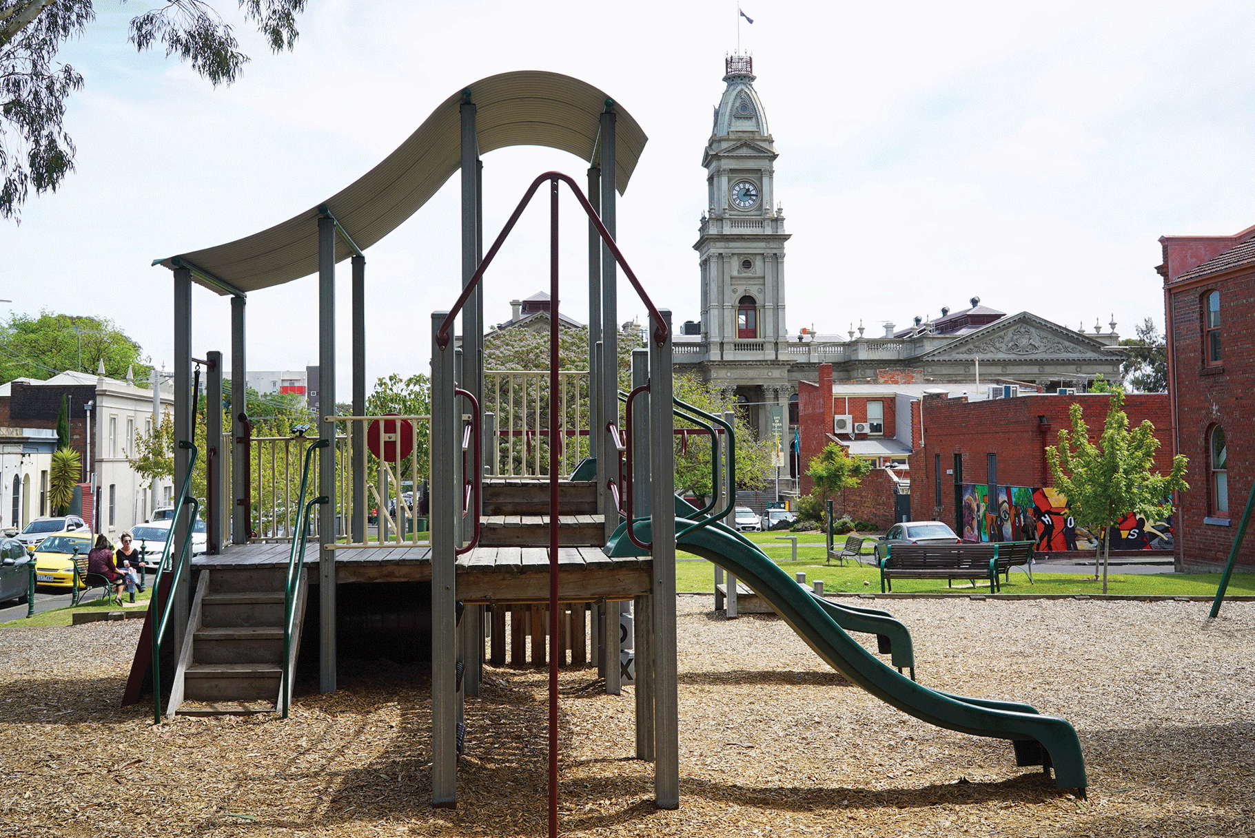 A playground with a wave shaped roof and a clock twoer in the background
