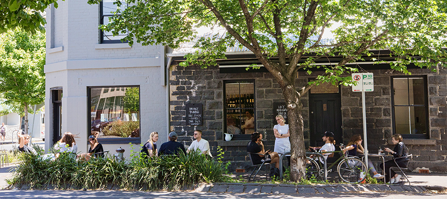 People sitting at tables and chairs on a footpath outside a hospitality business in Yarra, like a cafe or restaurant. The street is leafy and there are lots of parked bicycles.