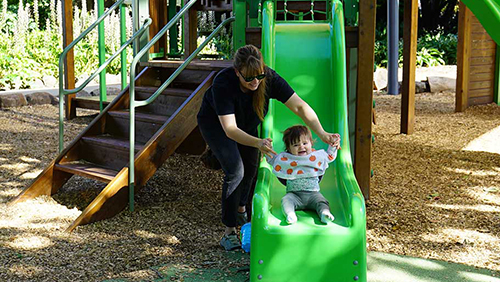 Mother helping toddler down playground slide