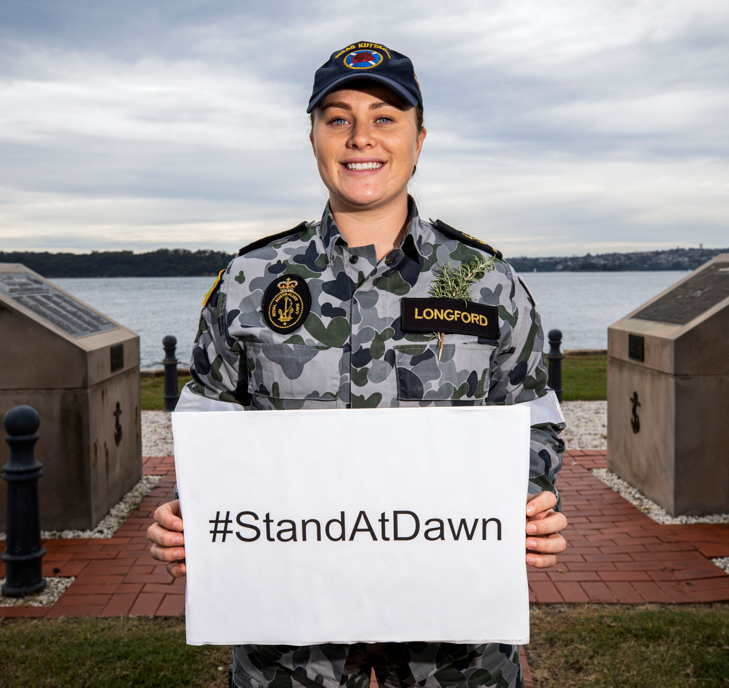 A female member of the Defence Force is by the bay holding a white sign with black text that reads #StandAtDawn