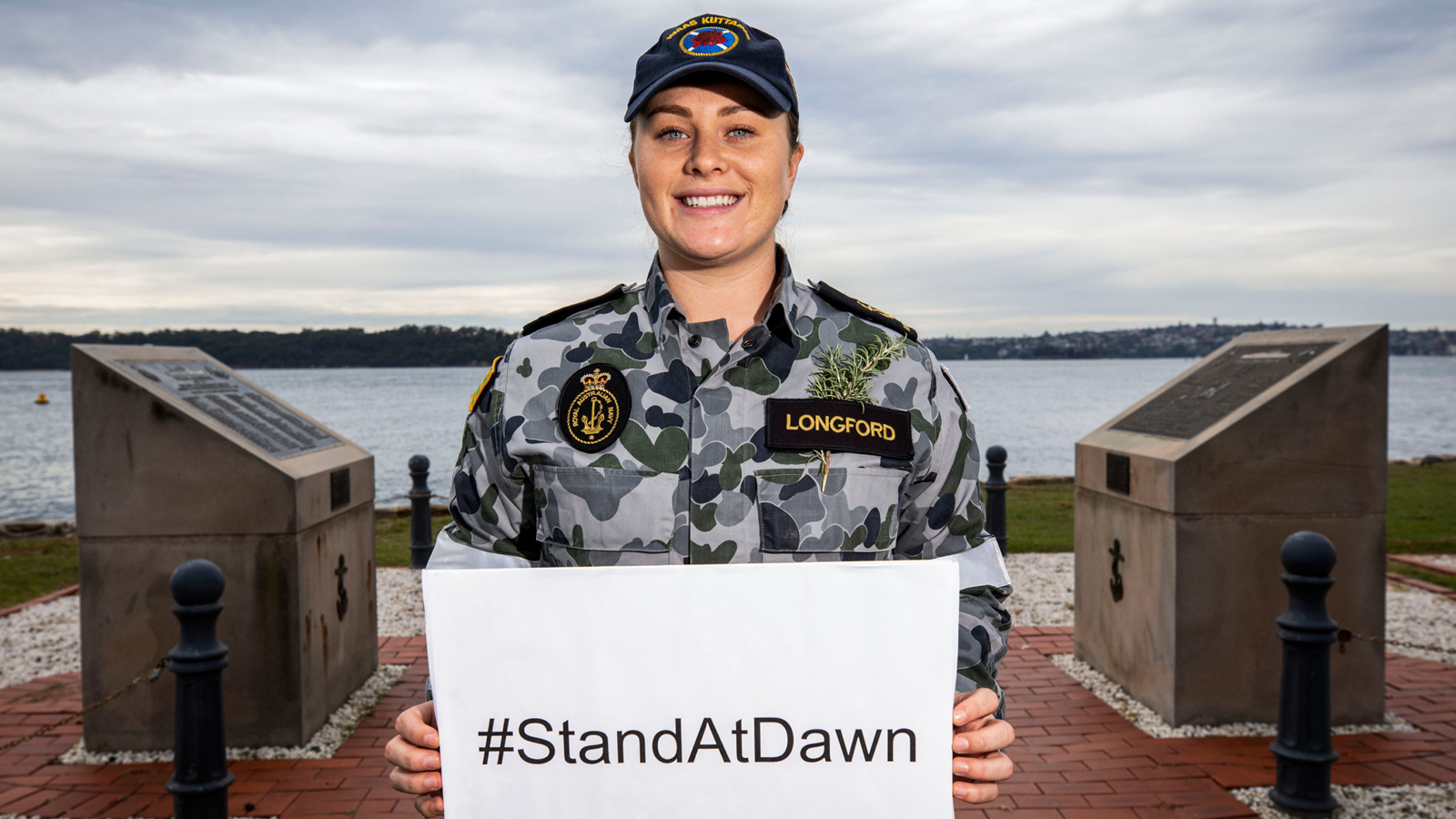 A female member of the Defence Force is by the bay holding a white sign with black text that reads #StandAtDawn