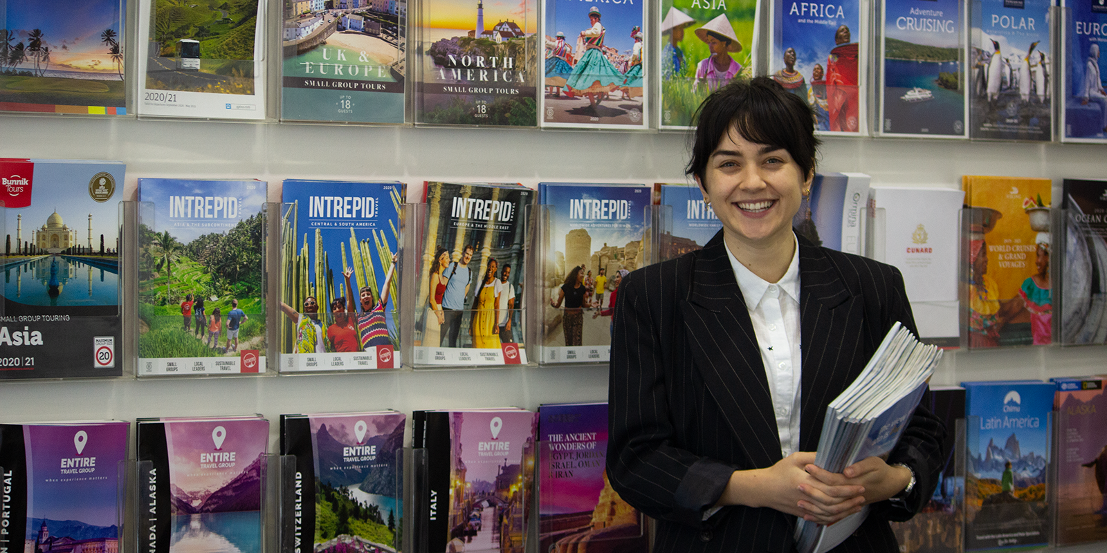 travel agent holding travel catalogues in front of a wall of magazines