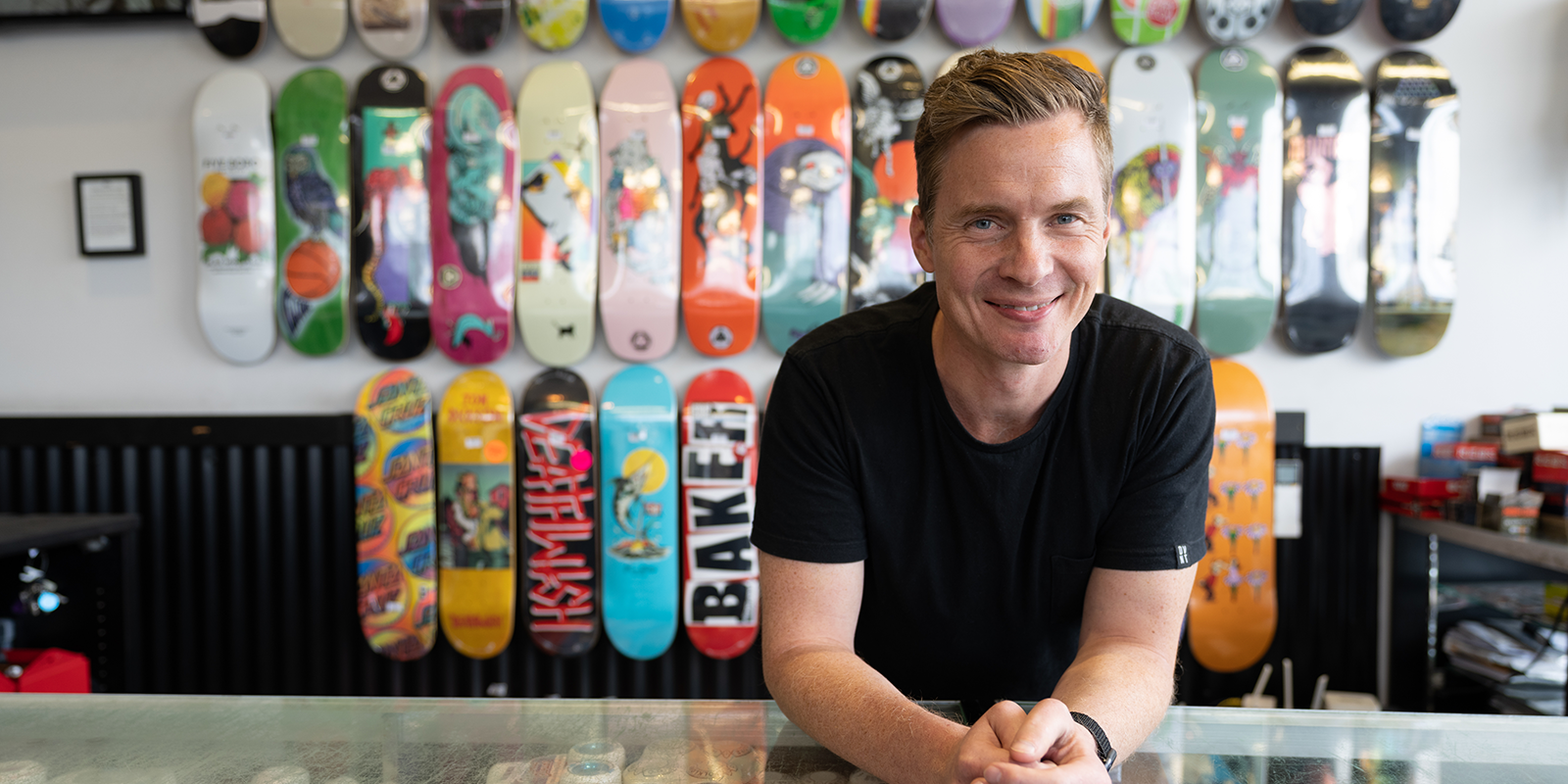 local trader leaning on glass cabinet with wall of skateboards behind him