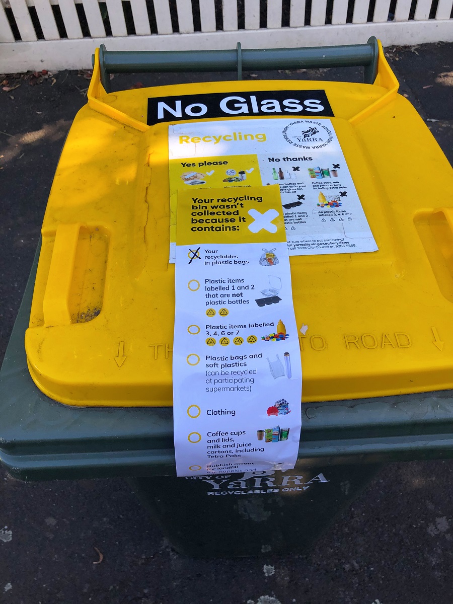 The lid of a yellow recycling bin with a sticker over it that lists incorrect bin items