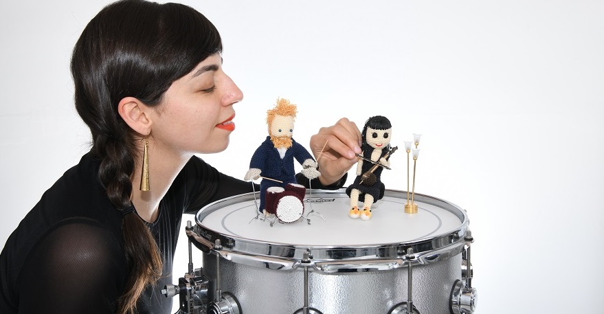 Woman with dark hair and gold earrings sitting next to a drum kit with two small toy figurines on top of it.  