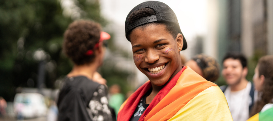 Young man smiling, looking into camera with rainbow flag wrapped around shoulders. People in background on street