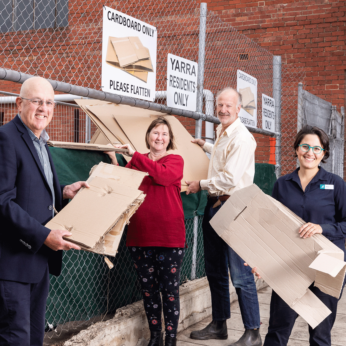 Four people holding flattened cardboard stand in front of bins and signs for cardboard recycling