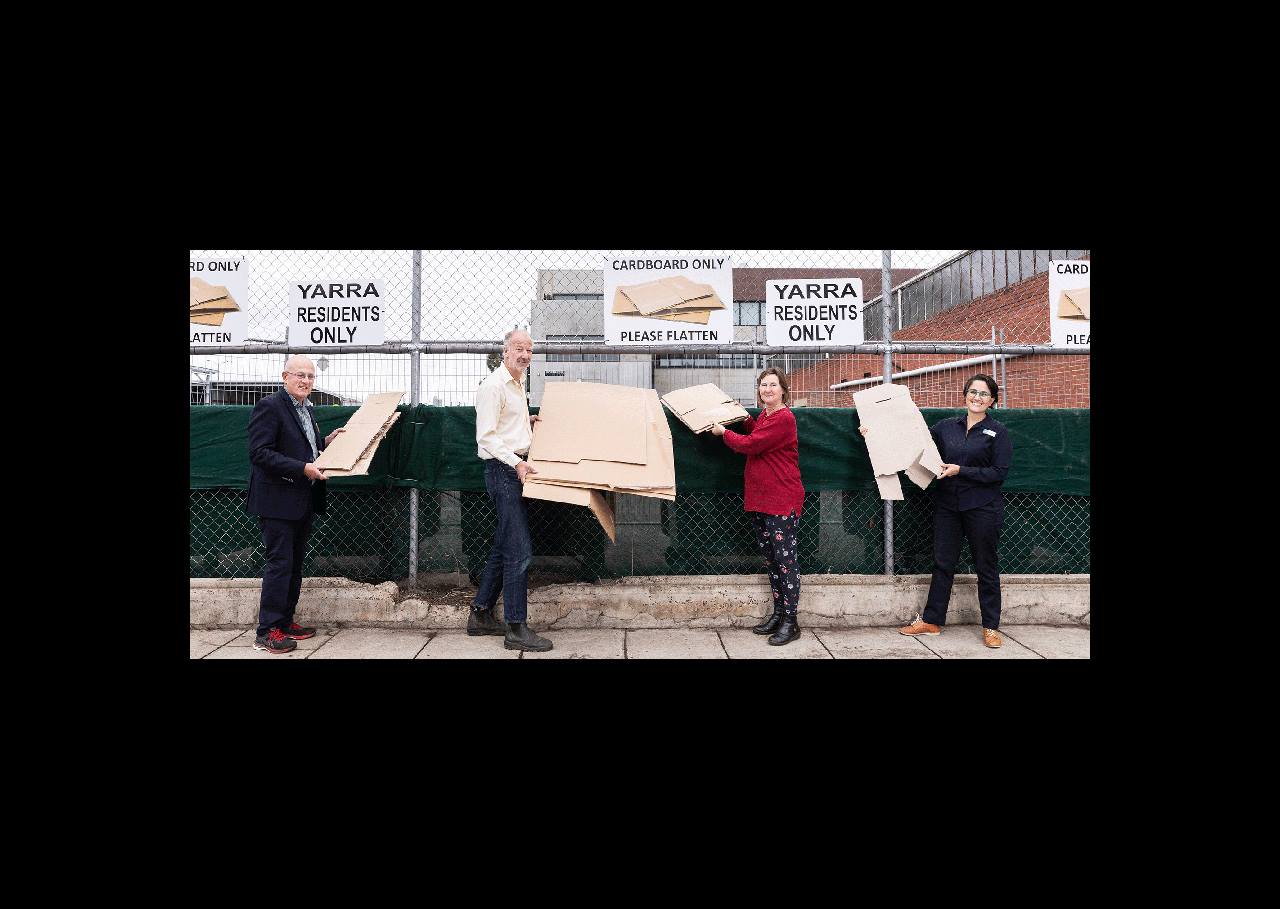 Four people holding cardboard stand in front of bins and a fence with signs for cardboard recycling