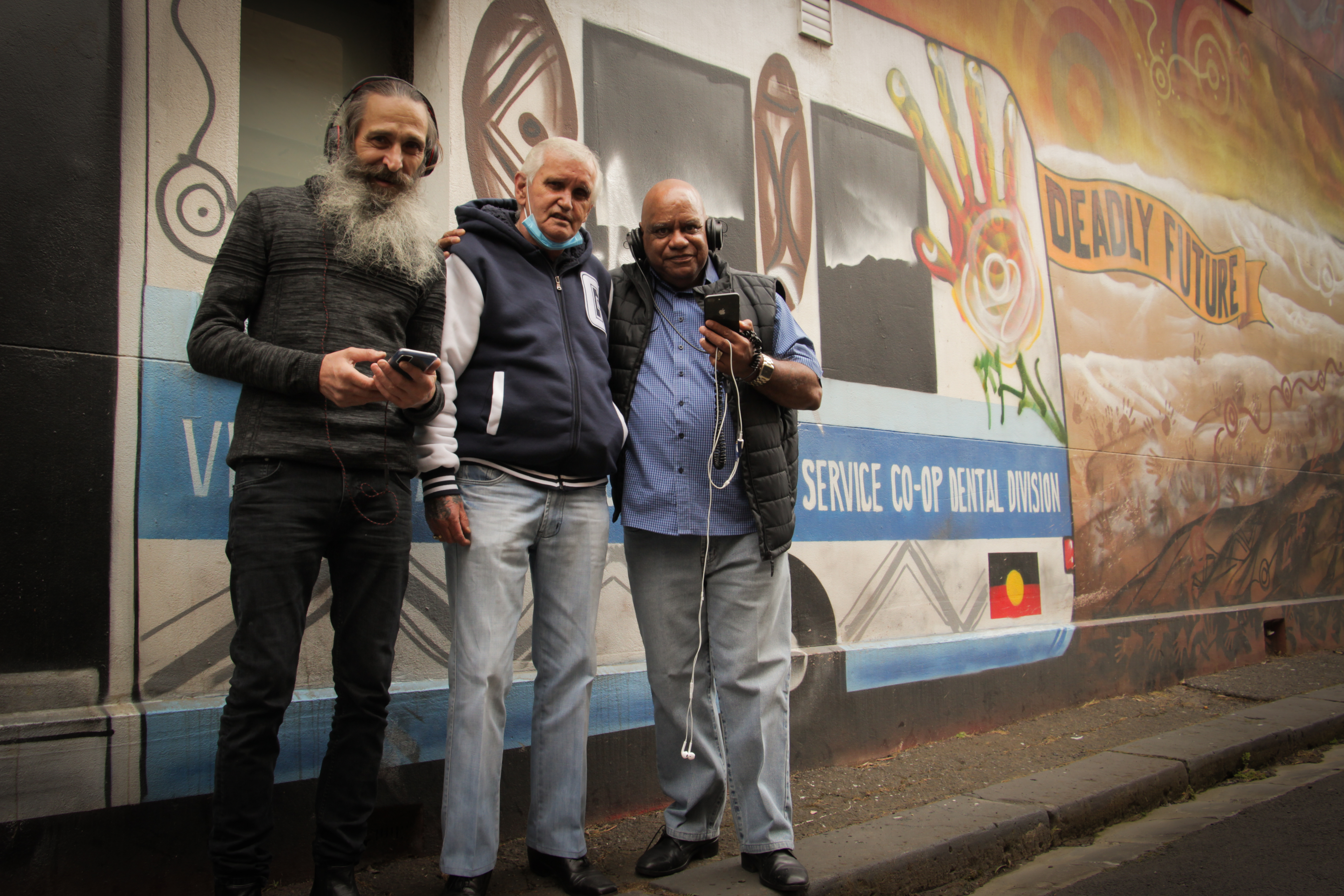 Uncle Colin Hunter, Uncle Bootsie Thorpe and elder Bobby Nicholls standing next to a mural that reads 'Deadly Future'. They are smiling, wearing headphones and holding their phones.