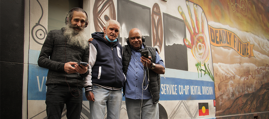 Uncle Colin Hunter, Uncle Bootsie Thorpe and Elder Bobby Nicholls standing and smiling, facing the camera in front of a mural that features the Aboriginal flag, 'Deadly Future' and other Indigenous artwork.