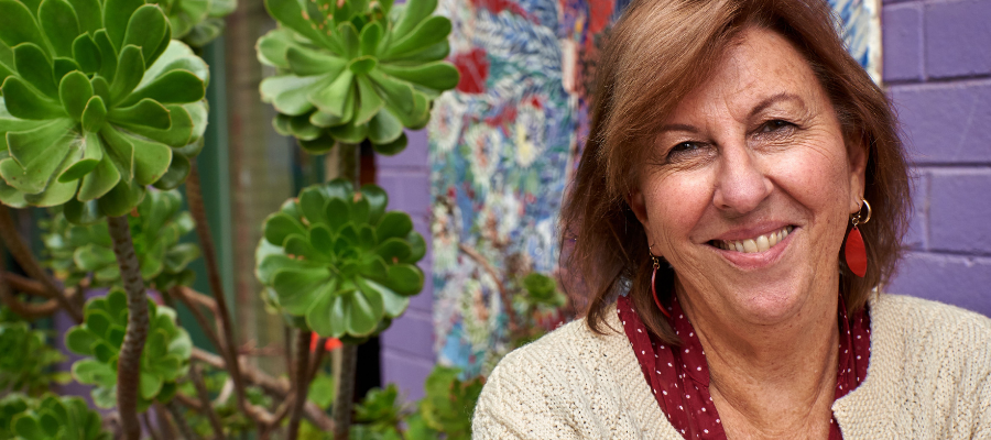 Woman in cream cardigan, standing in front of purple wall and plant smiling into camera