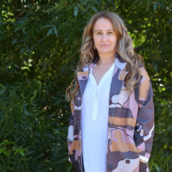 Woman standing in front of green foliage, wearing white shirt and colourful patterned jacket 