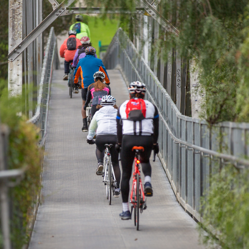 Cyclists wearing helmets and backpacks, cycling in a line away from camera across bridge