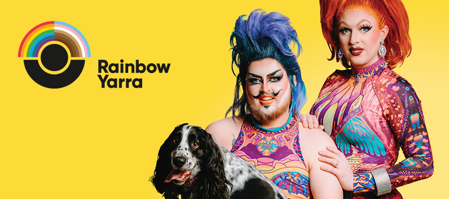 Two drag artists smiling for a photo with a dog against a yellow background with the Rainbow Yarra logo.