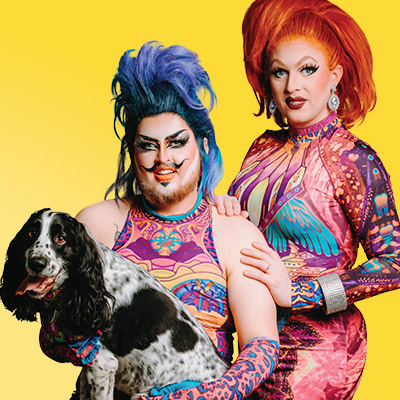 Two drag artists smiling for a photo with a dog against a yellow background.