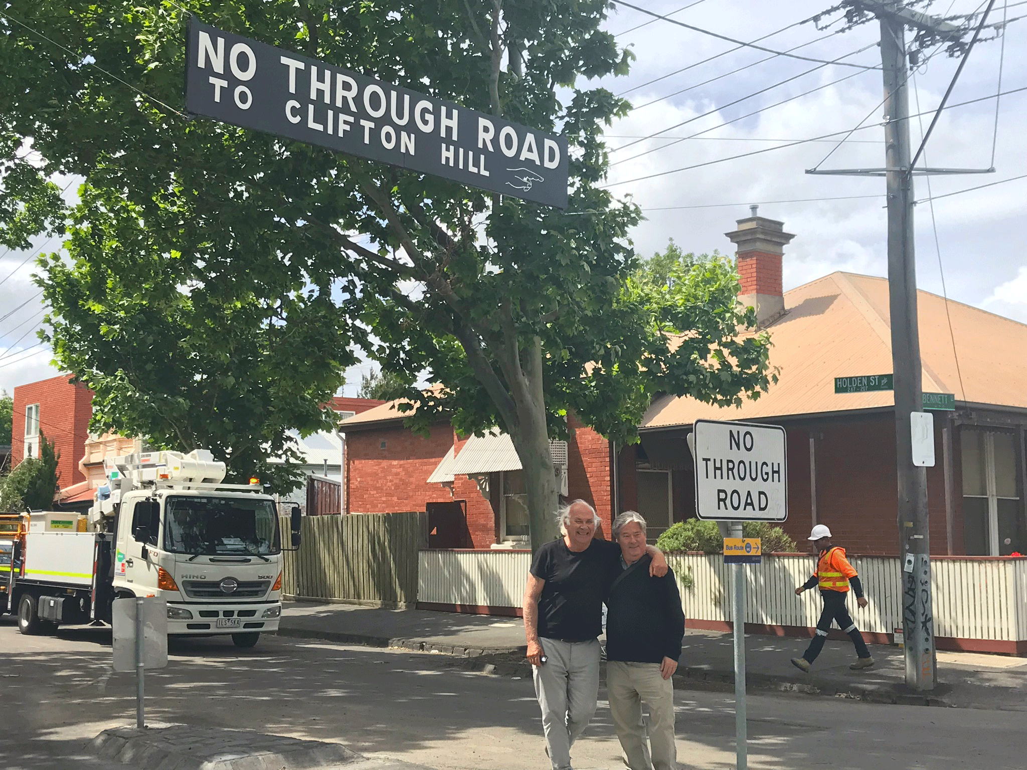 Two men standing arm in arm beneath a sign which says No Through Road