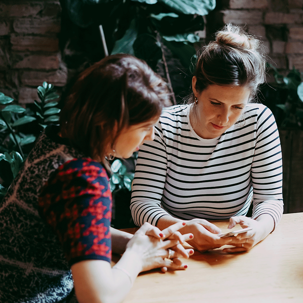 Two women sit in a green leafy cafe with serious expressions discussing an app on a phone