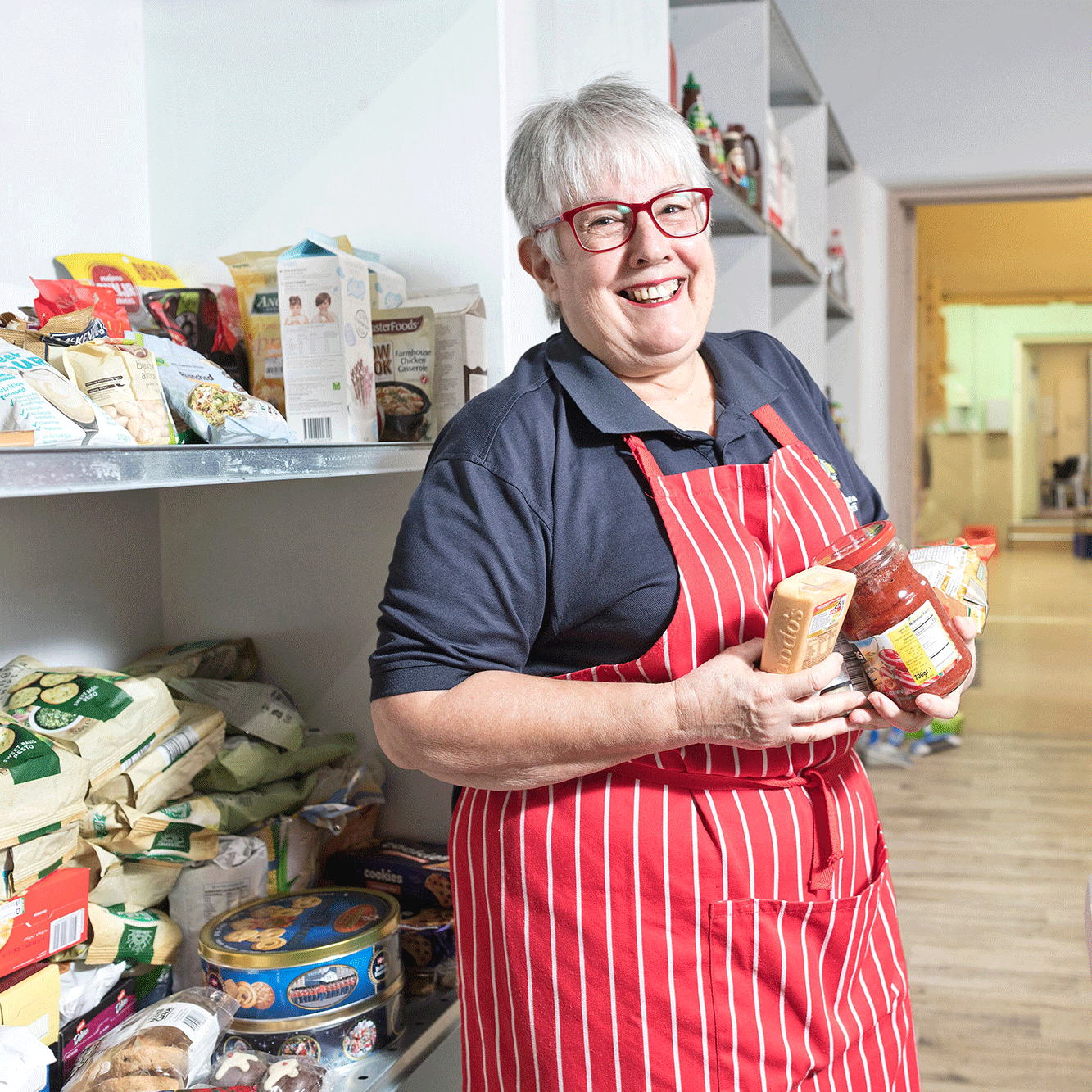 A smiling woman in a red apron surrounded by donated food