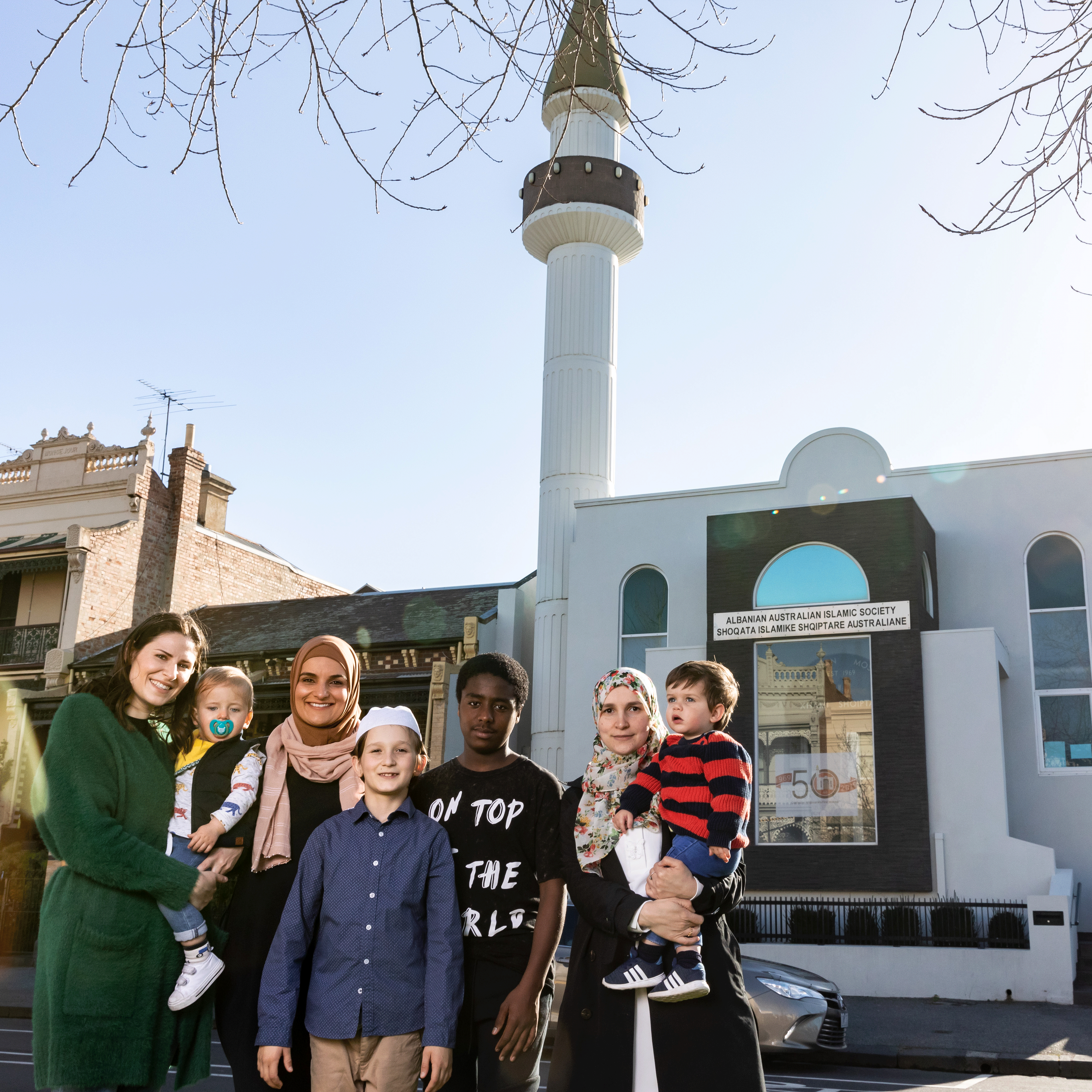 A group of women, young people and children stand outside in the sunshine in front of a white mosque 