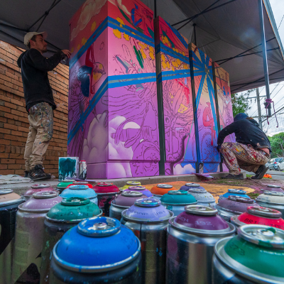 2 artists spray painting community battery in purple and pinks, depicting birds and plants