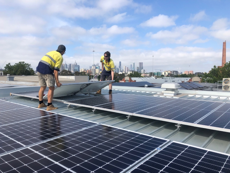 Two people wearing hi-vis installing solar panels on a roof. Melbourne city skyline in the background