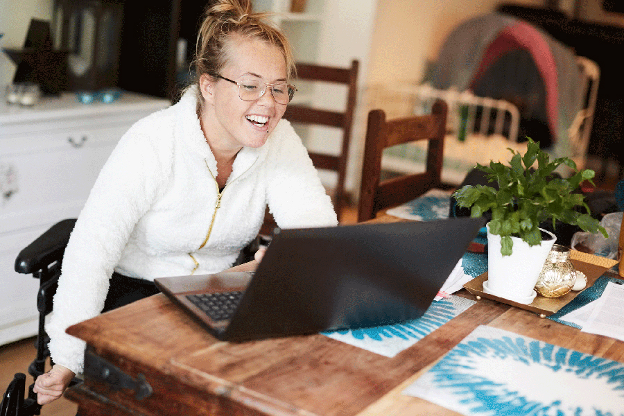 A woman smiling into her laptop at her kitchen table. She is sitting in a wheelchair. 