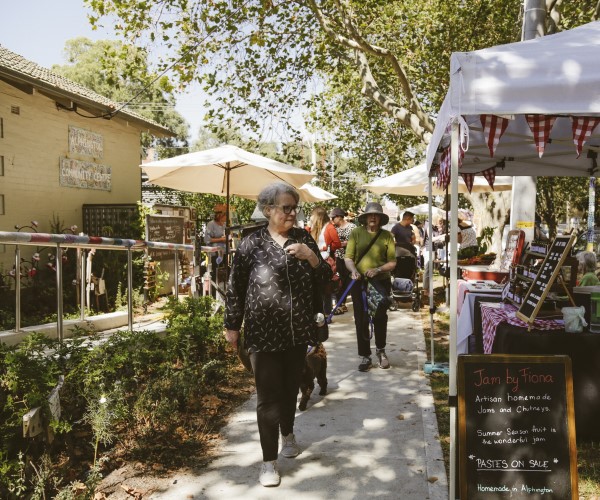 An outdoor market with stalls and umbrellas. Tall trees in background. Yellow building on left side.
