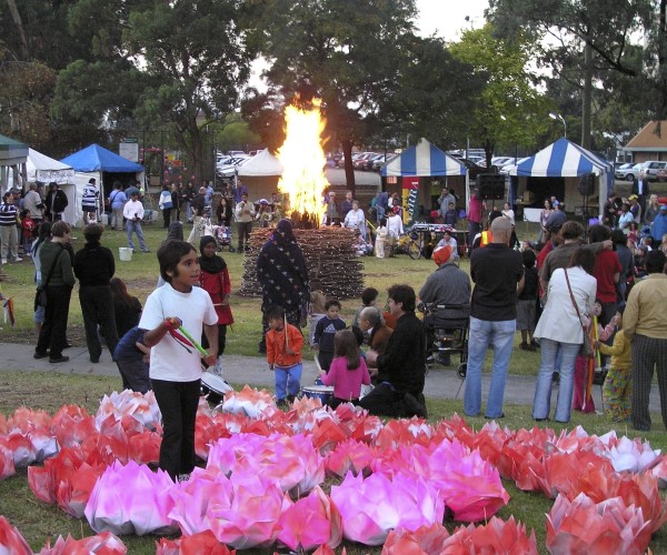 Outdoor event. Child standing in pink flowers in foreground and large bonfire in background.