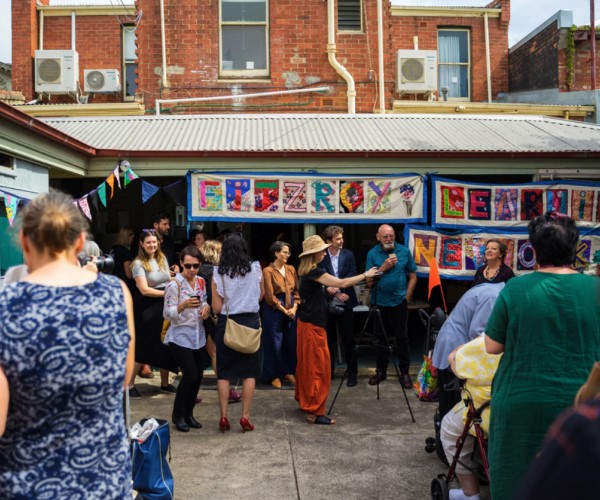 Group of people standing outside a house. A colourful stitched quilt hangs from the verandah roof that says 'Fitzroy Learning Network'
