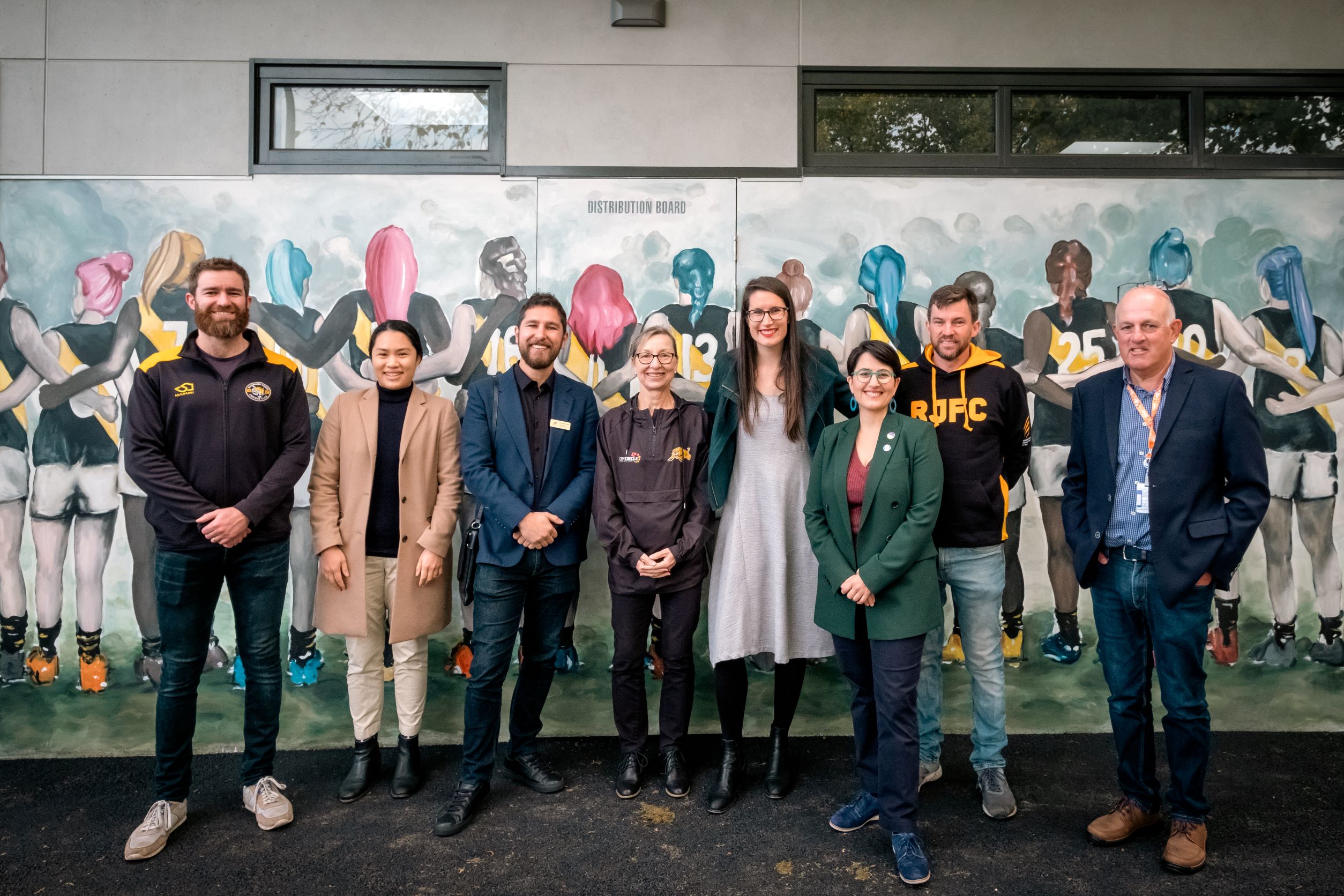 A group of men and women representing Richmond sporting clubs and Yarra City Council stand in front of a mural that pictures a women's football team standing arm in arm with their backs turned. 