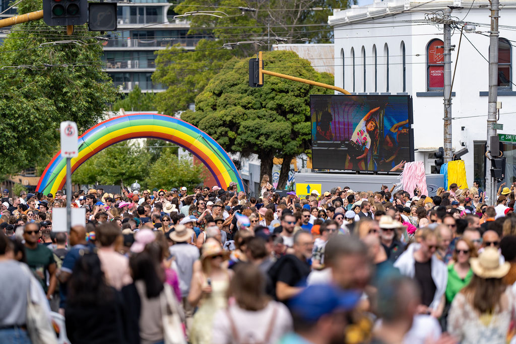 A crowd of people fill a street at Victoria's Pride Street Party in Fitzroy. A blow-up rainbow is in the background, and a person on a large television screen can be seen dancing.
